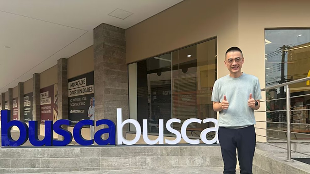 Alex Ye fazendo joinha ao lado do letreiro Busca Busca na entrada da loja